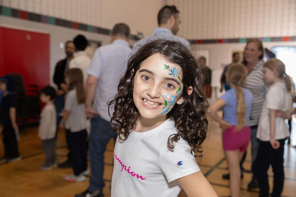 Young girl with curly brown hair and flower face paint smiles for a photo